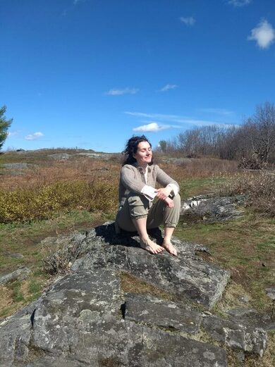 Victoria Maria sits with her feet on a rock in a pasture framed by a bright blue sky with a few small white clouds. Her eyes are closed.